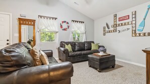A living room with a brown leather couch, a smaller couch, and a wooden coffee table. There is a window with white curtains and a wreath on the wall. - Welcome to “Whimsy Woods Cabin” – The Ultimate Cabin Retreat on Lake Cumberland (Burnside)