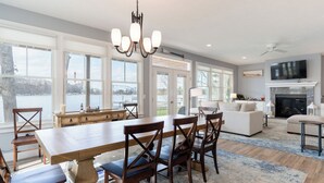 A spacious dining area with a long wooden table and chairs, a chandelier, and large windows overlooking a body of water.