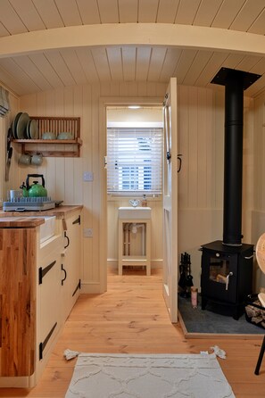 A wooden interior with a kitchen area, a black stove, and a window with blinds. - Shepherd's Hut Near Cotswolds & Sky Dive Fun (Brackley)