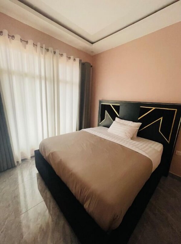Relax in a stylish room with a large bed, beige comforter, and white pillows, surrounded by a light peach wall and large window.
