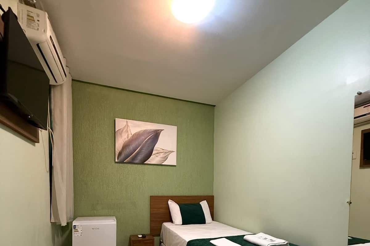 A single-bed room with a green wall, a ceiling light, a small refrigerator, a bedside table, and a mirror.