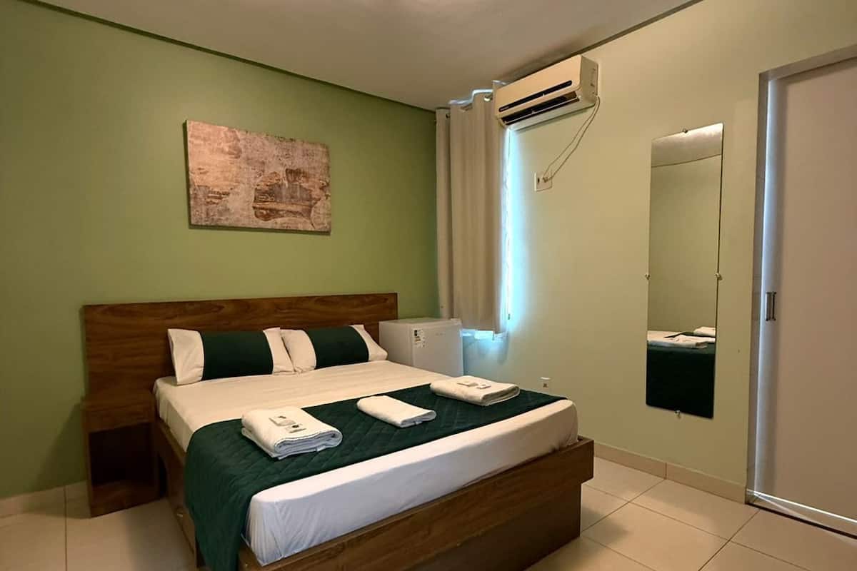 A bedroom with a wooden bed, green bedding, a wall-mounted air conditioner, a mirror, and a painting on the wall.