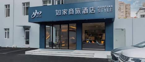 A hotel entrance with a modern facade, a clear glass door, and a parking area with a white car.