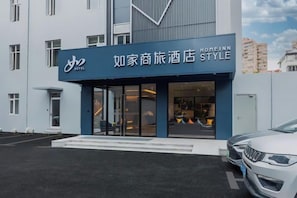 A hotel entrance with a modern facade, a clear glass door, and a parking area with a white car.