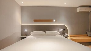 Een moderne hotelkamer met een groot bed, een nachtkastje met lamp en een zithoekje met kussen.