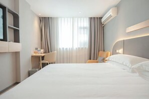 A hotel room with a large bed, a desk with a chair, a TV mounted on the wall, and an air conditioning unit.