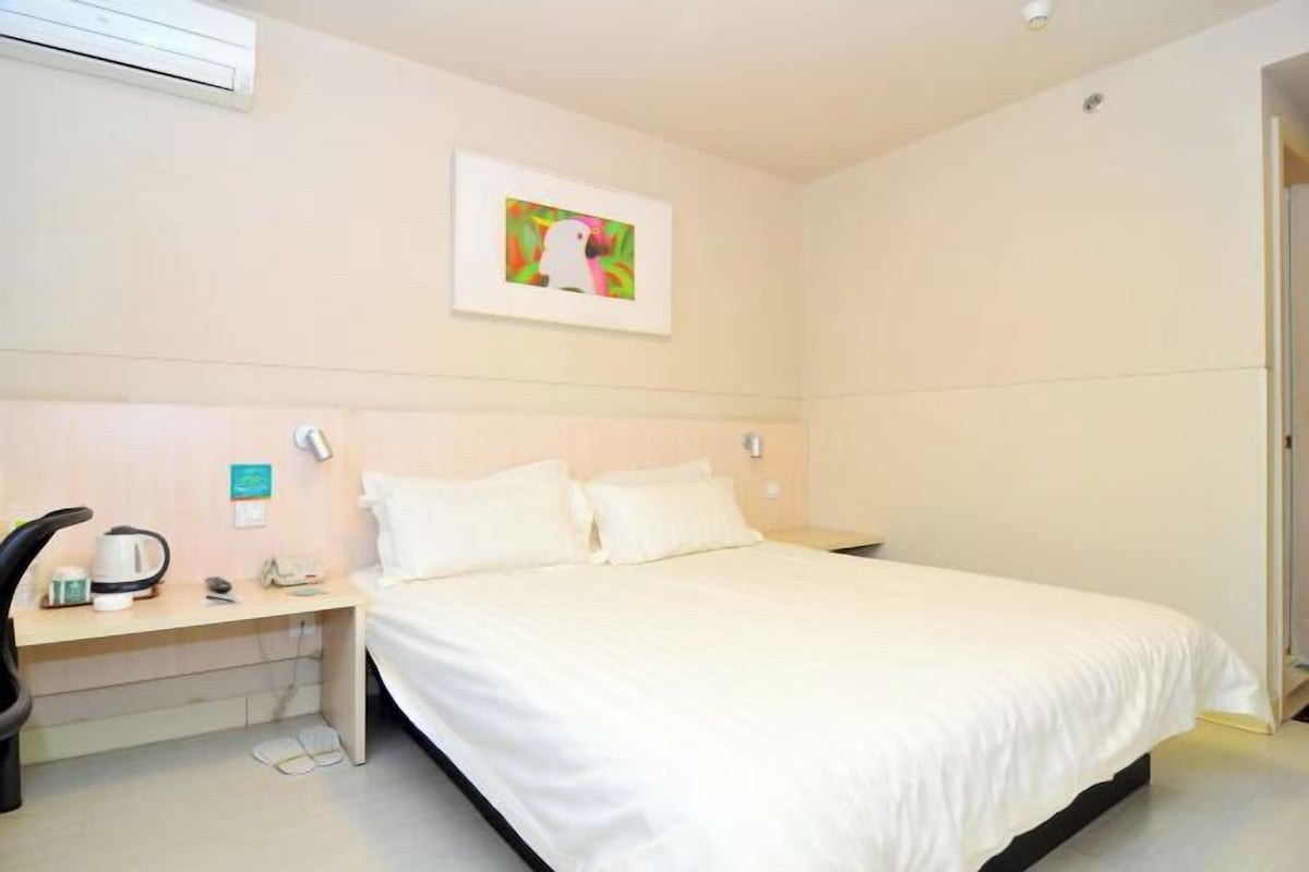 A hotel room with a bed, a desk with a kettle, a chair, and a picture on the wall.