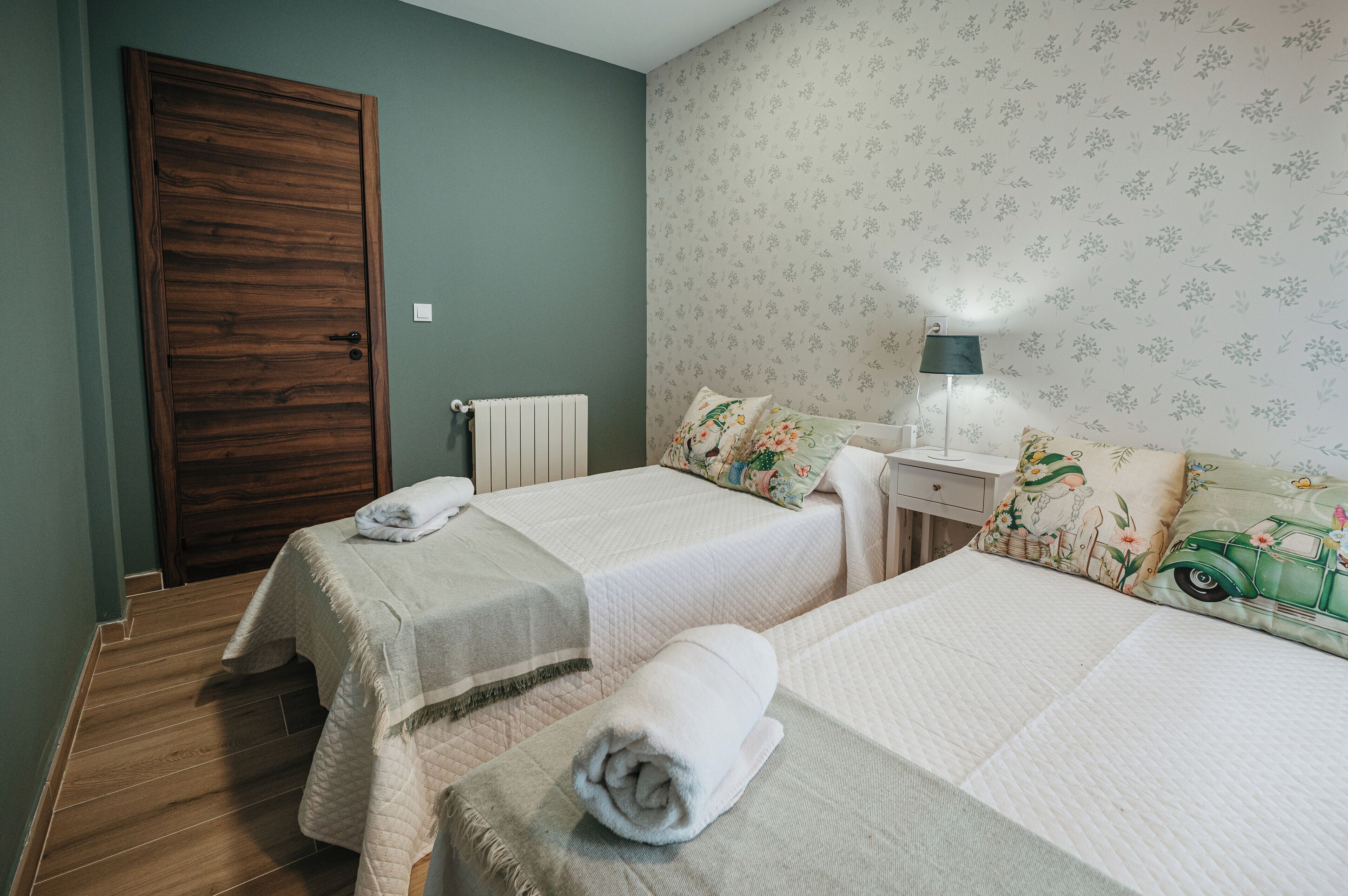 Una habitación con dos camas, una puerta de madera, un radiador y una mesita de noche con lámpara.