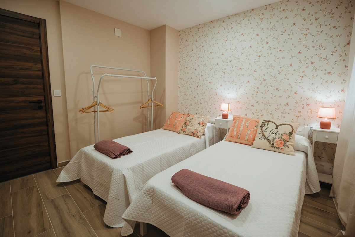 Hotellihuone, jossa on kaksi sänkyä, puuovi, seinälle kiinnitetty pyyheteline ja kukallinen tapetti.