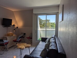 Een moderne woonkamer met een bank, stoelen, een televisie en een groot raam met uitzicht op bomen.