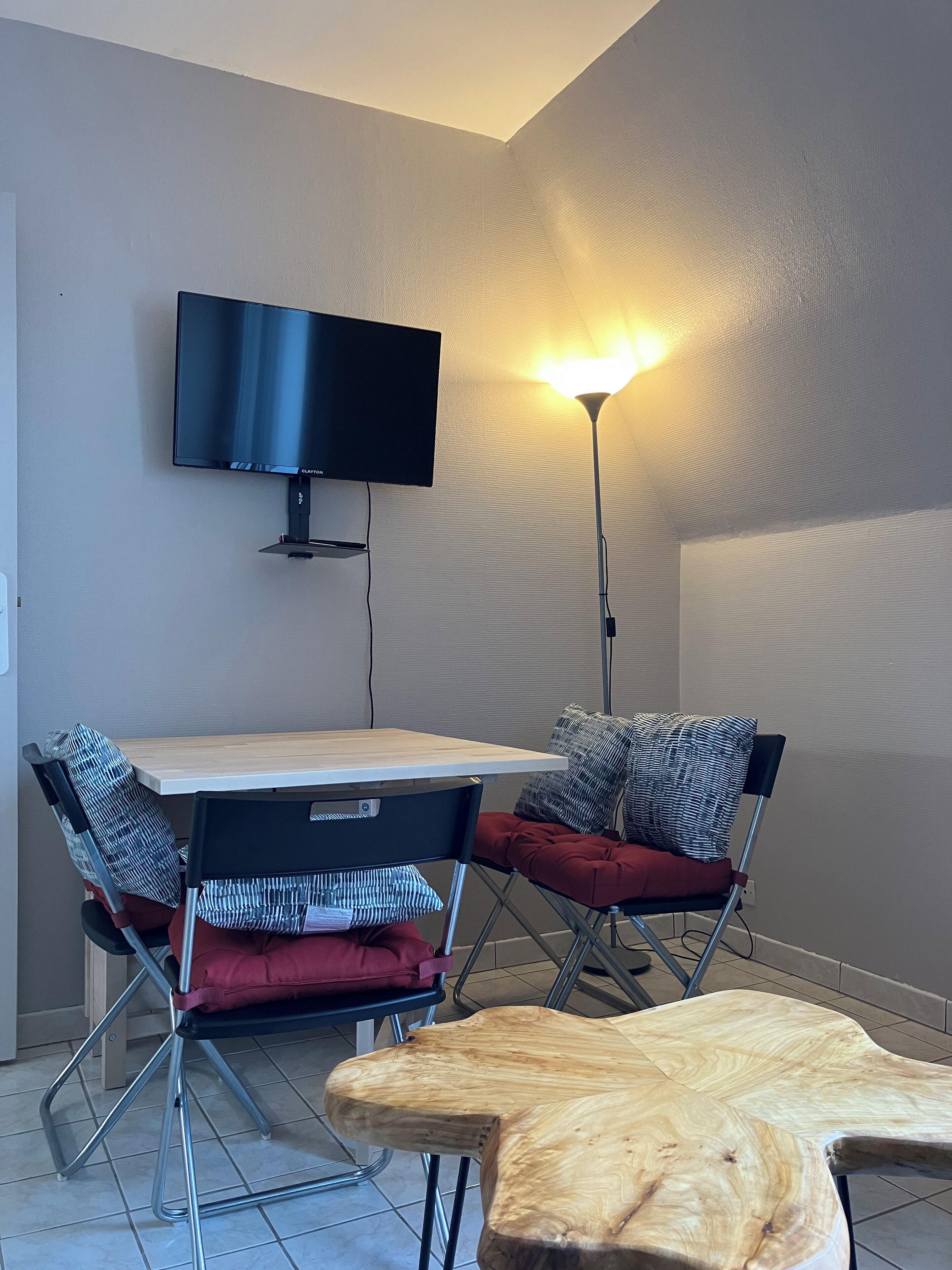 Una habitación moderna con una televisión de pantalla plana montada en la pared, una mesa de madera, dos sillas con cojines rojos y una lámpara de pie.
