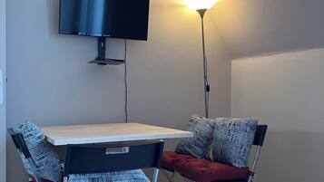 Una habitación moderna con una televisión de pantalla plana montada en la pared, una mesa de madera, dos sillas con cojines rojos y una lámpara de pie.