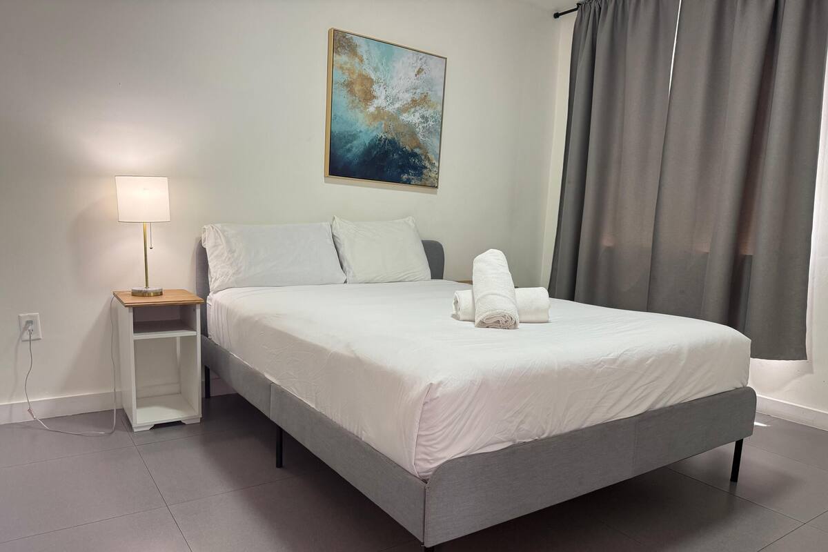Een net opgemaakt bed met witte lakens, een nachtkastje met een lamp en een schilderij aan de muur.