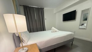Habitación de hotel con una cama, una mesita de noche con lámpara, un televisor montado en la pared y un pequeño cuadro enmarcado.