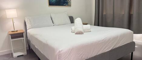 A neatly made bed with white linens and two rolled towels, a bedside table with a lamp, and a painting on the wall.