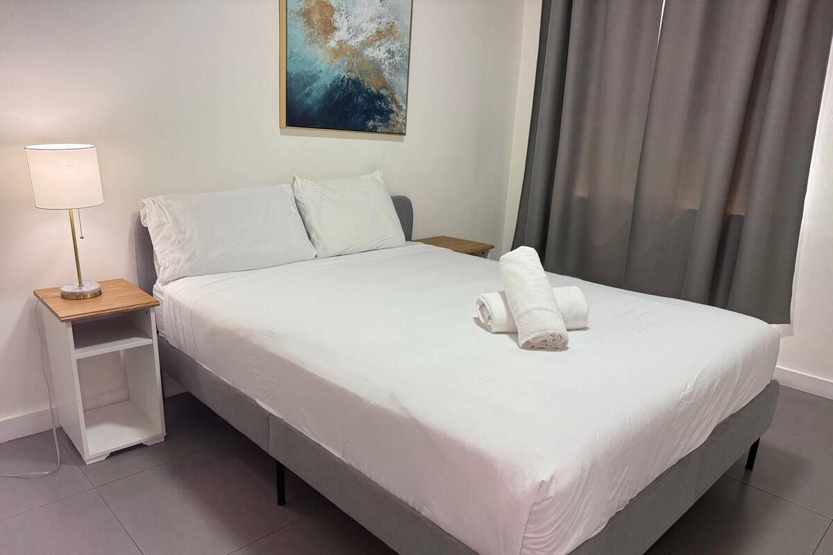 Een netjes opgemaakt bed met witte lakens en twee opgerolde handdoeken, een nachtkastje met een lamp, en een schilderij aan de muur.