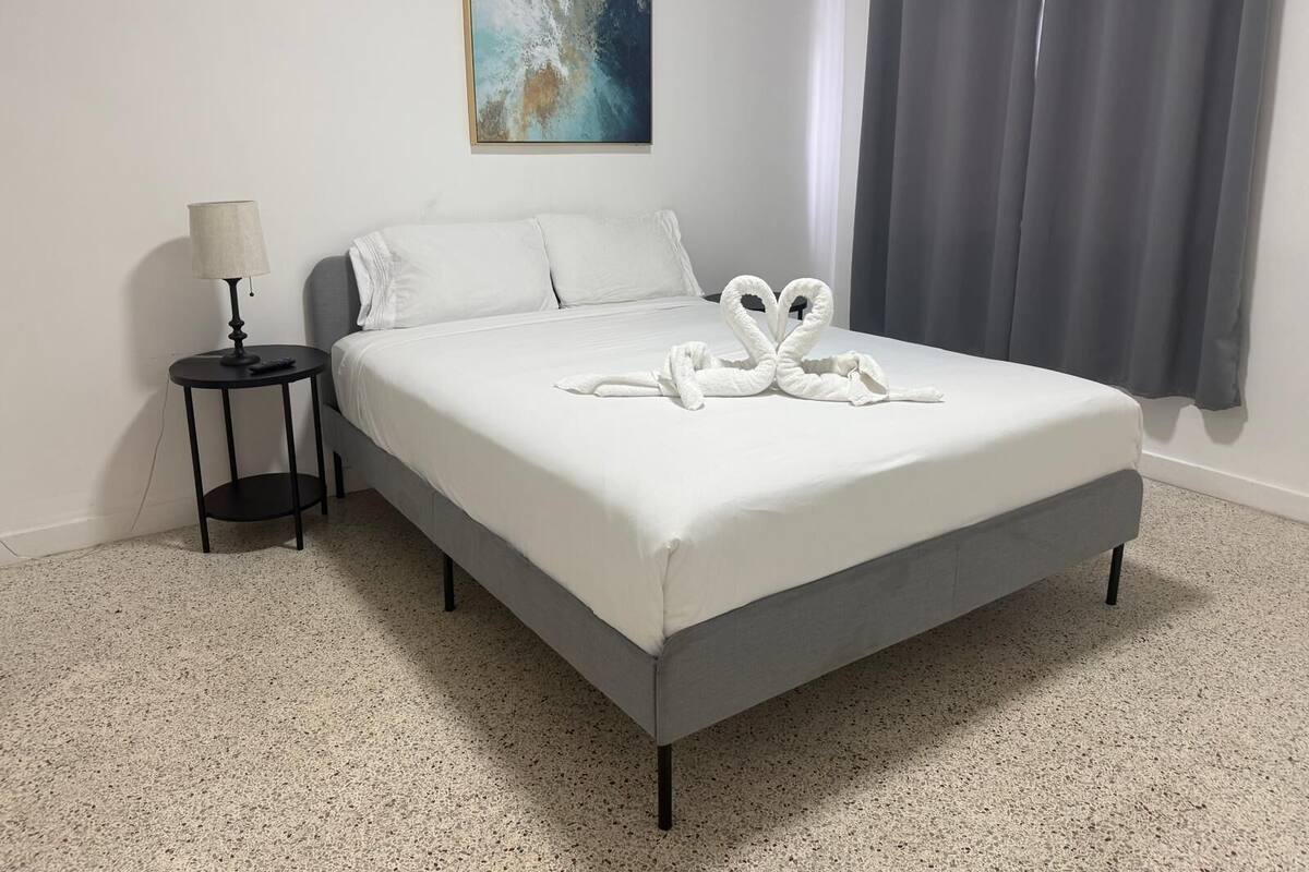 Een netjes opgemaakt bed met een handdoek in de vorm van een zwaan, een klein nachtkastje met een lamp en een schilderij aan de muur.