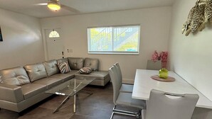 A modern living room with a sectional sofa, a glass coffee table, a dining table with chairs, and a large window with a view of a staircase. - Stylish 1-bed apartment, in East Delray Beach, pets, parking, free laundry (Delray Beach)