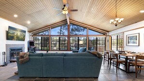 A spacious living area with a large sofa, dining table, and chairs, a fireplace, and large windows offering a view of the outdoors.