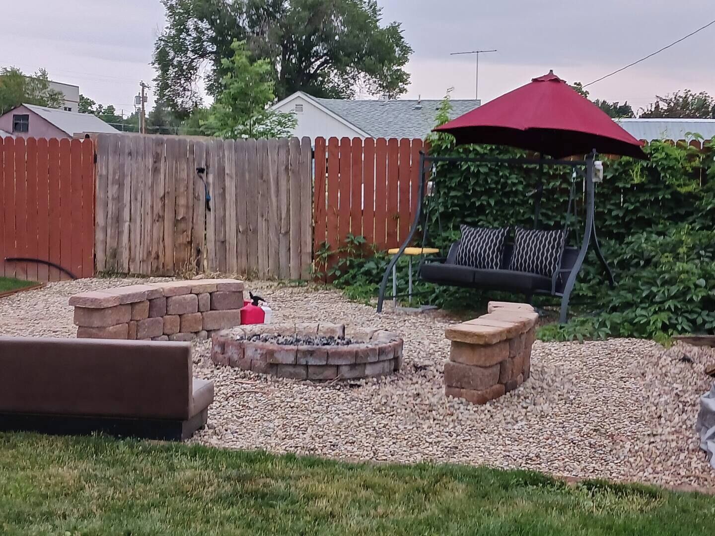 A backyard with a wooden fence, a red umbrella, a fire pit, and a stone bench.
