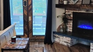 Uma sala de estar acolhedora com um banco de madeira, uma mesa de vidro com uma bebida, uma lareira e uma vista através de uma porta.