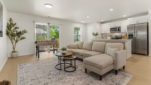 Ein modernes Wohnzimmer mit einem beigefarbenen Ecksofa, einem Couchtisch und einem Teppich. Es gibt einen Esstisch mit Stühlen, eine Küchenzeile mit weißen Schränken und Geräten sowie ein großes Fenster mit Jalousien.