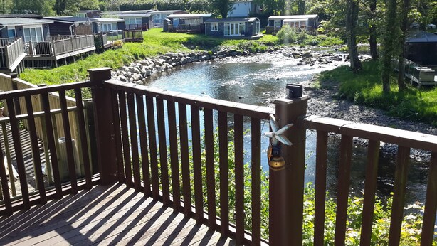 A wooden deck overlooking a river with a view of mobile homes in the background. - Cosy holiday chalet (Cearnarfo)