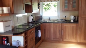 A kitchen with wooden cabinets, a stove, a sink, and a window with a view of greenery. - Cosy holiday chalet (Cearnarfo)