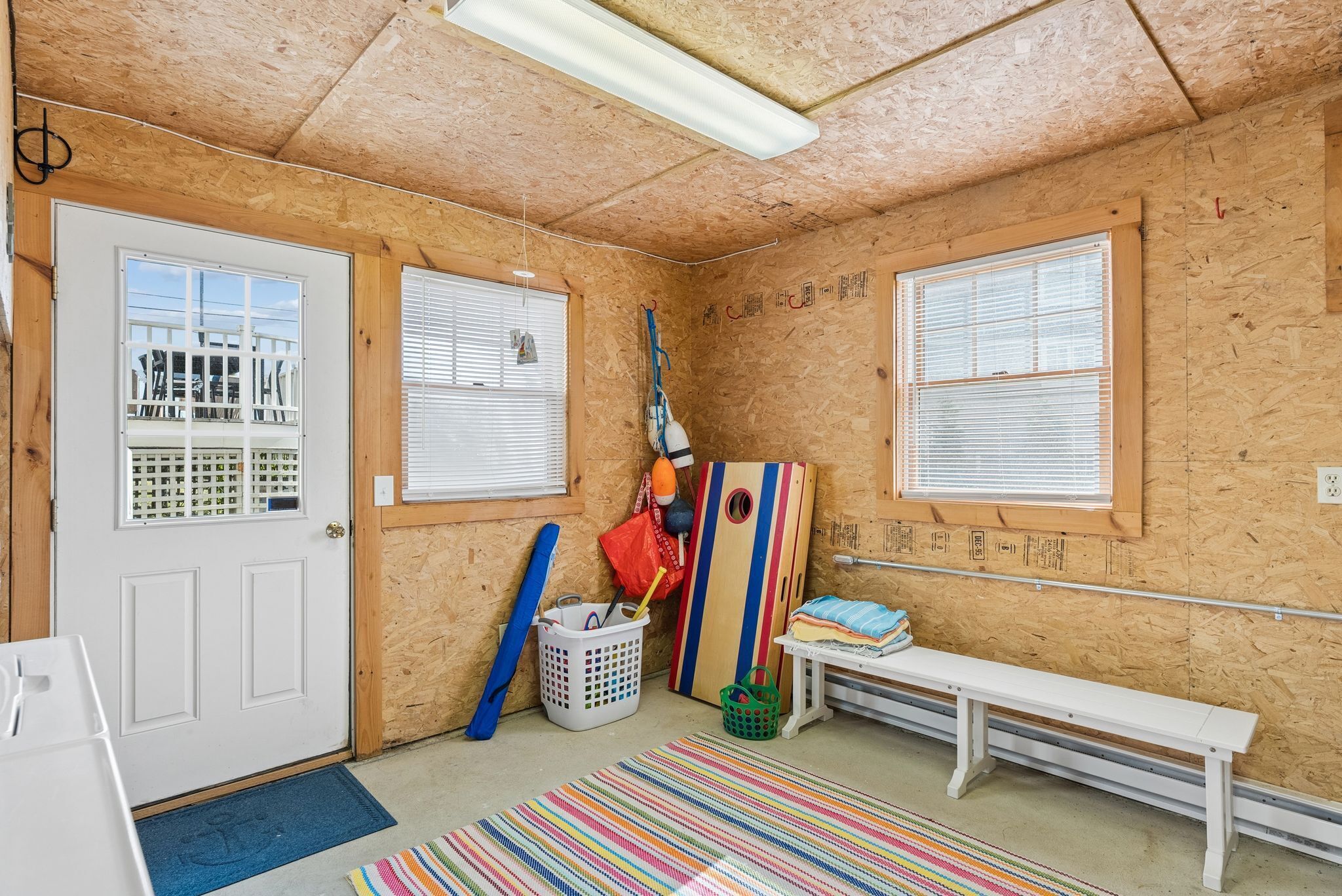 A room with wooden walls, two windows with blinds, a bench, a colorful striped rug, and a door with a window grille.