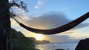 Un hamac tendu entre deux arbres, offrant une vue sur une plage rocheuse, l’océan et le coucher de soleil.
