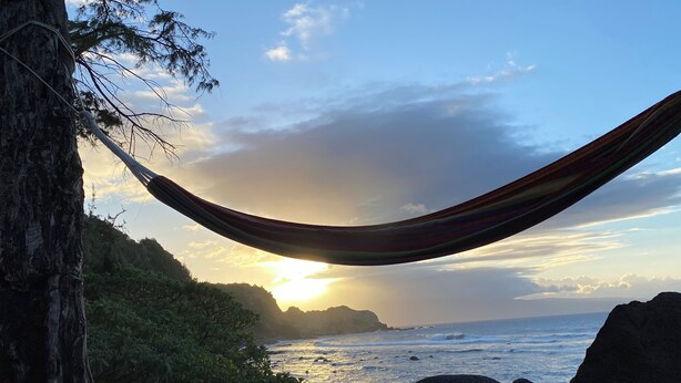 A hammock tied between trees overlooking a rocky beach with the ocean and sunset.