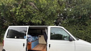 A white van with an open back, revealing a bed with a blue and white patterned bedspread and a wooden floor.