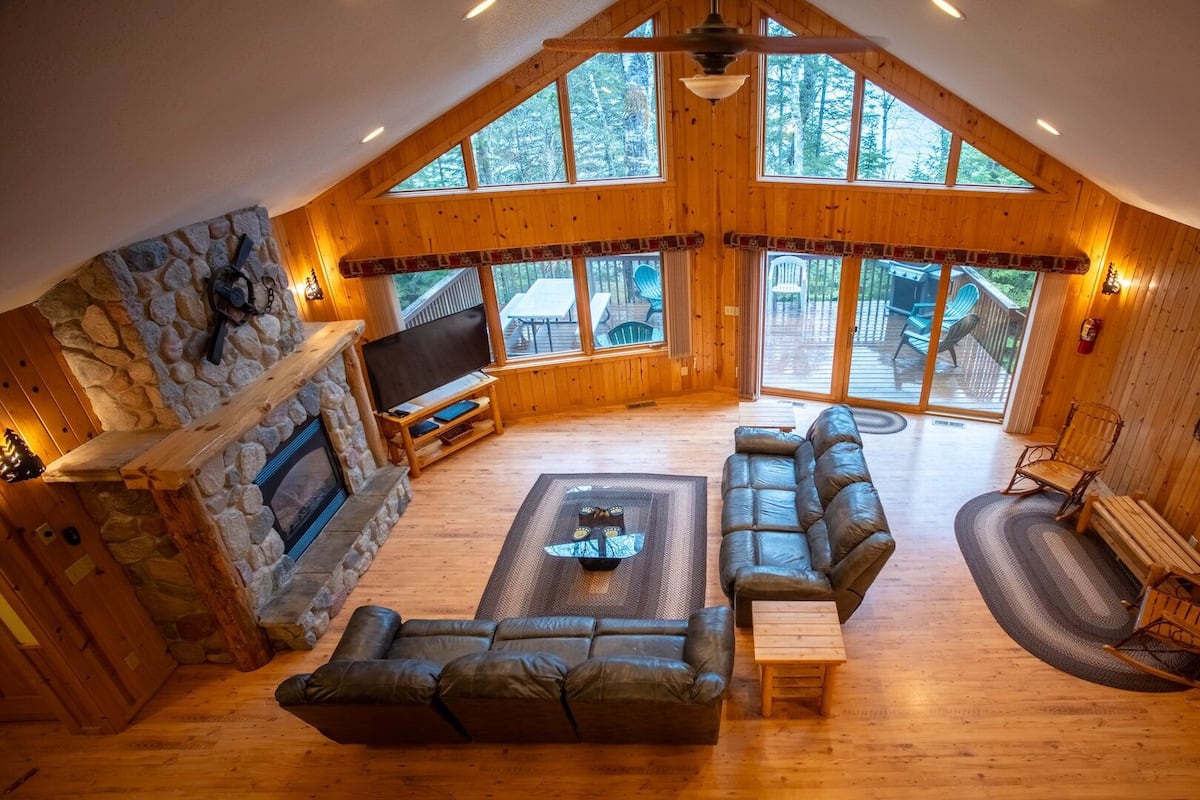 A cozy cabin interior with a stone fireplace, leather sofas, a TV, and large windows overlooking a deck.