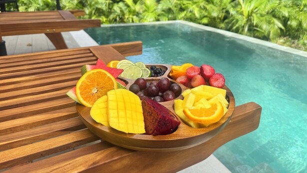 Et træbord med en frugtfat ved en pool, omgivet af frodig vegetation.