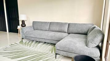 A grey sectional sofa with a side table and a lamp, a woven floor rug, and a round wicker side table.
