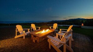 A fire pit table with four Adirondack chairs in a patio area with a view of the ocean and distant mountains.