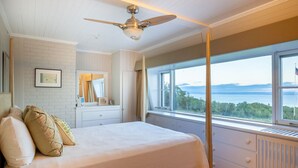 A bedroom with a bed, a ceiling fan, a window with a view of the sea, and a white brick wall.