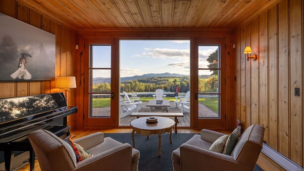 A cozy room with a piano, two armchairs, a round table, and a painting. Large windows offer a view of a patio with outdoor furniture.
