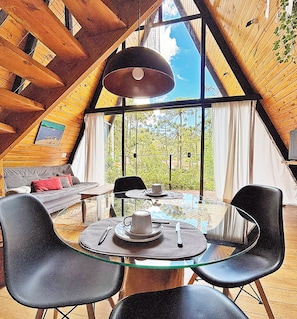 Un intérieur chaleureux avec une table en verre, des chaises noires, un canapé et une grande fenêtre donnant sur les arbres.