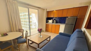 Un espacio habitable compacto con kitchenette, una zona de comedor con mesa y sillas, un sofá, y vistas a un balcón con un edificio al fondo.