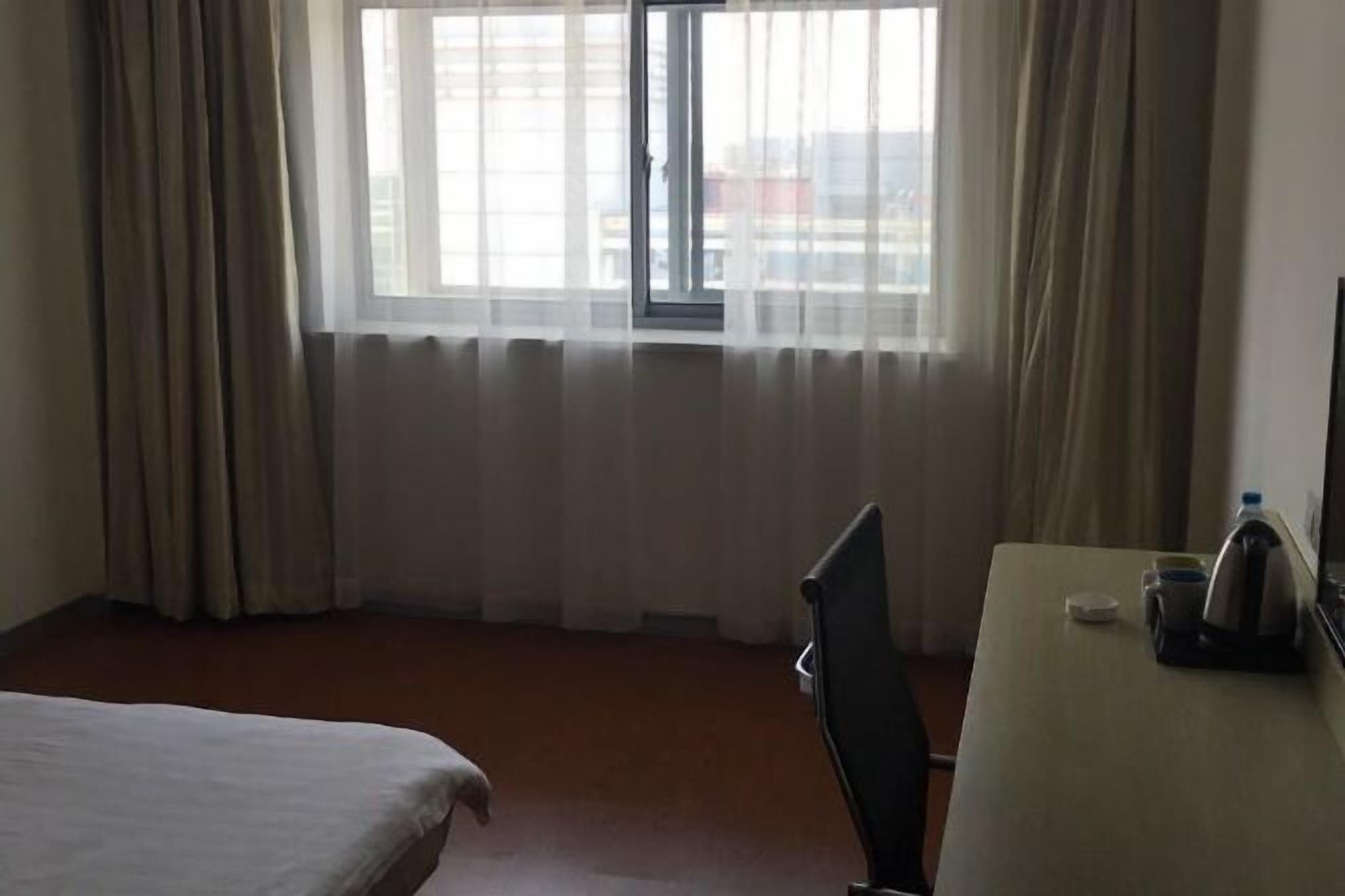 A hotel room with a bed, a desk, a chair, a window with curtains, and a view of buildings outside.