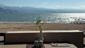 Een tafel met een bloevas, zout- en peperstel en bestek op een balkon met uitzicht op een strand en bergen.