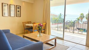 Hotelkamer met een blauwe bank, een eettafel met stoelen, een vaas met bloemen en een balkon met uitzicht op zee en palmbomen.