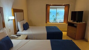 Habitación de hotel con dos camas, televisor, escritorio con teléfono y ventana con vista a la playa y palmeras.