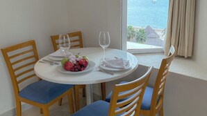 A dining table with a fruit plate, wine glasses, and cutlery, set against a window with a view of the sea and a thatched-roof structure.