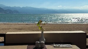 A table with a vase of flowers, salt and pepper shakers, and cutlery on a balcony overlooking a beach with mountains in the background. - Oceanfront: Views and Amenities! (Puerto Vallarta)
