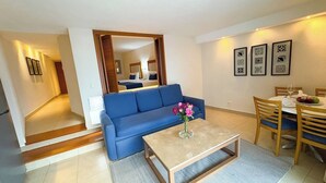Ein modernes Wohnzimmer mit einem blauen Sofa, einem Couchtisch mit Blumenvase, einem Essbereich mit Tisch und Stühlen und einem Flur, der zum Schlafzimmer führt.