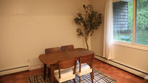 A dining area with a wooden table and chairs, a small plant, and a window with a view of greenery.
