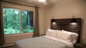 A bedroom with a large window, a bed with white bedding, a wooden headboard, and a nightstand.