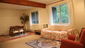 A cozy living room with a wooden ceiling, a sofa, a red armchair, a dining table with chairs, and a large window with curtains.